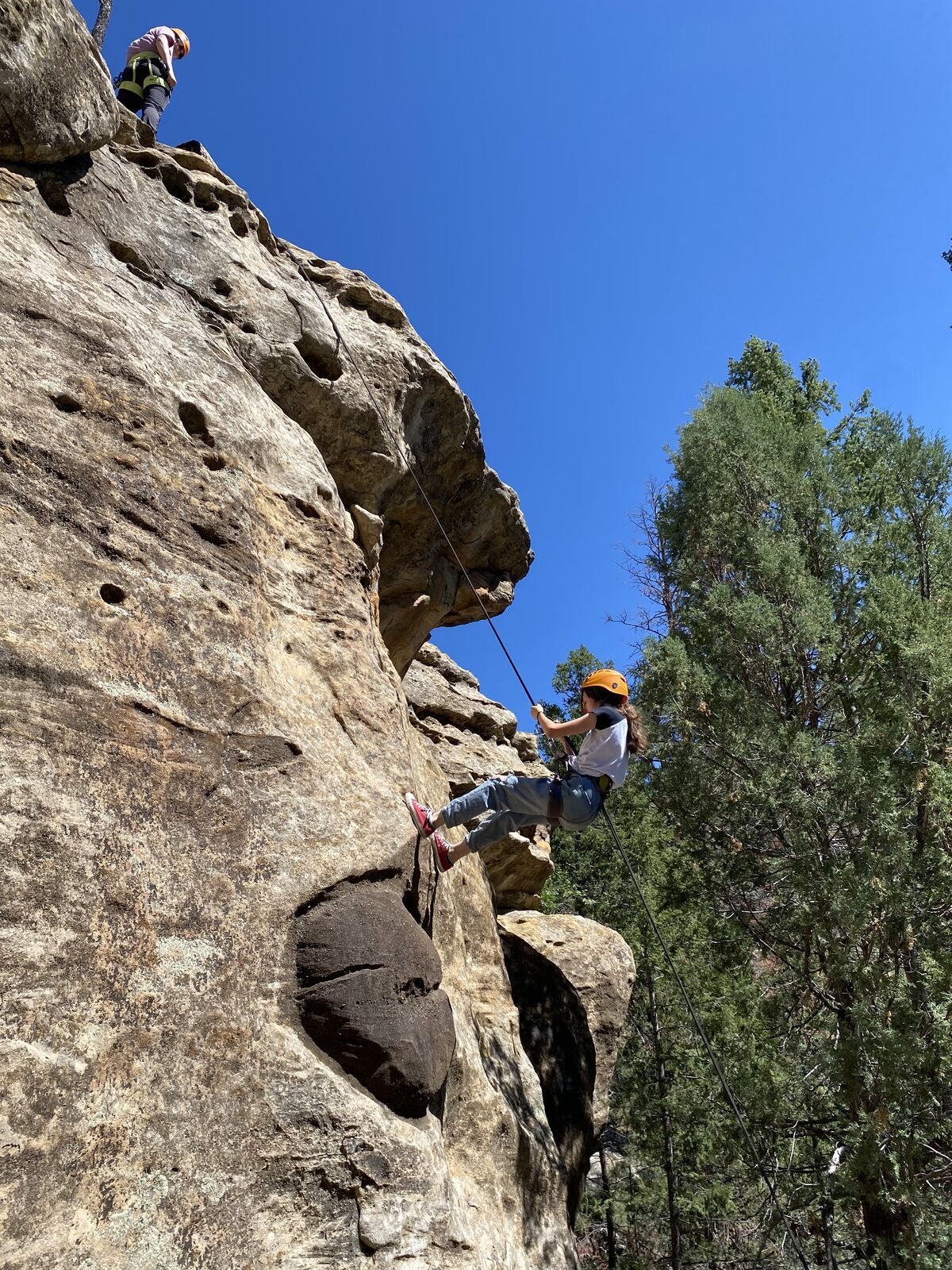 A climber in an orange helmet lowers down a pocketed sandstone face against a deep blue sky.