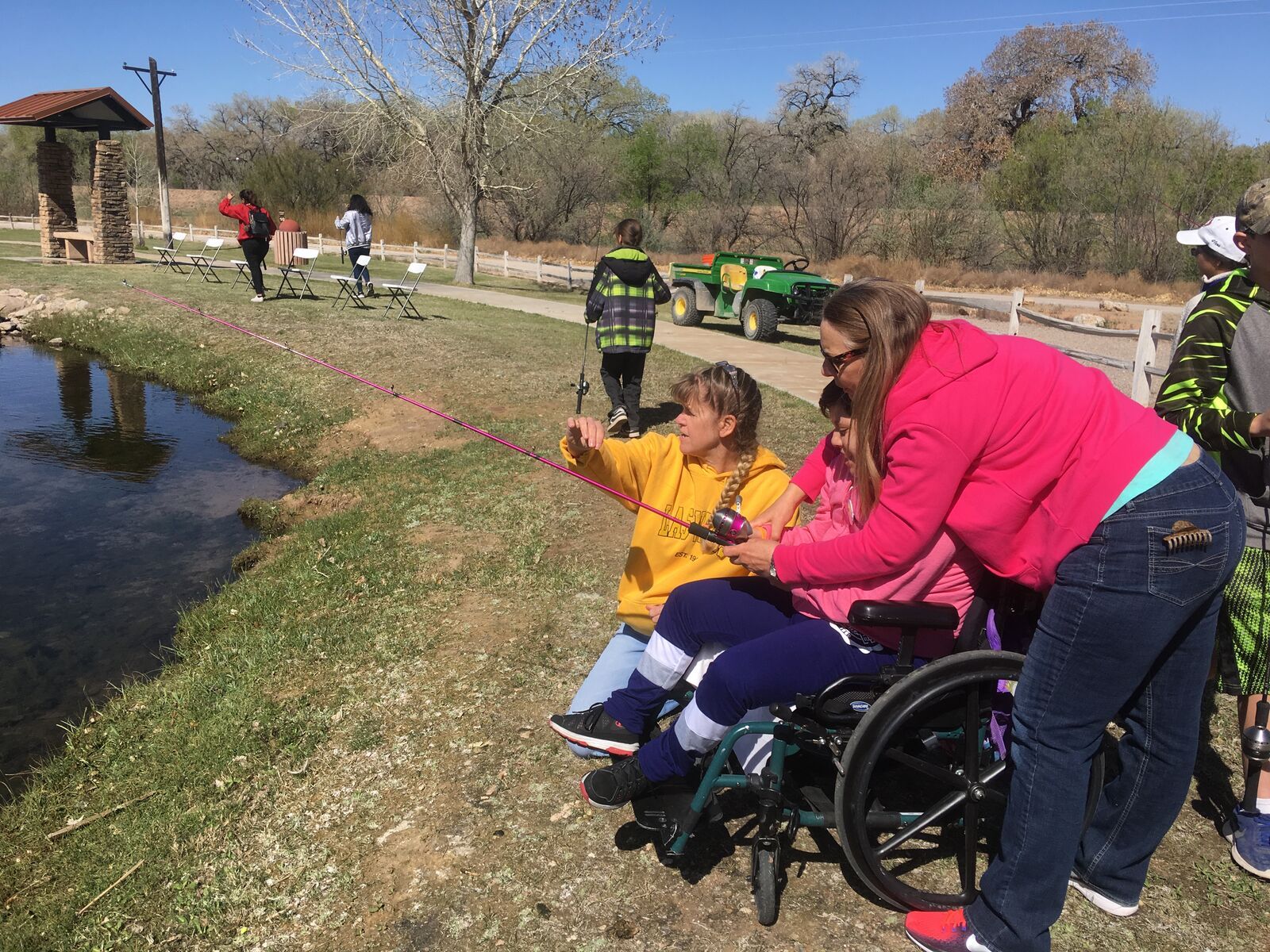 A child in a wheelchair fishes from a grassy bank with help from a volunteer.