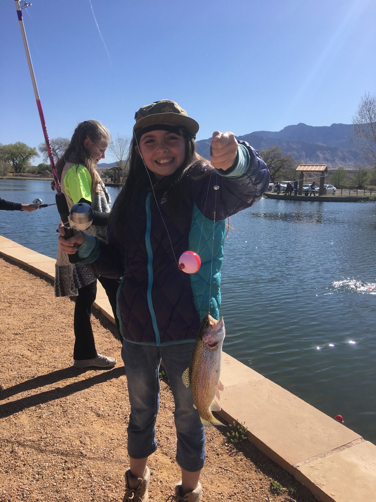 A girl holds up a trout on a line beside a pond.