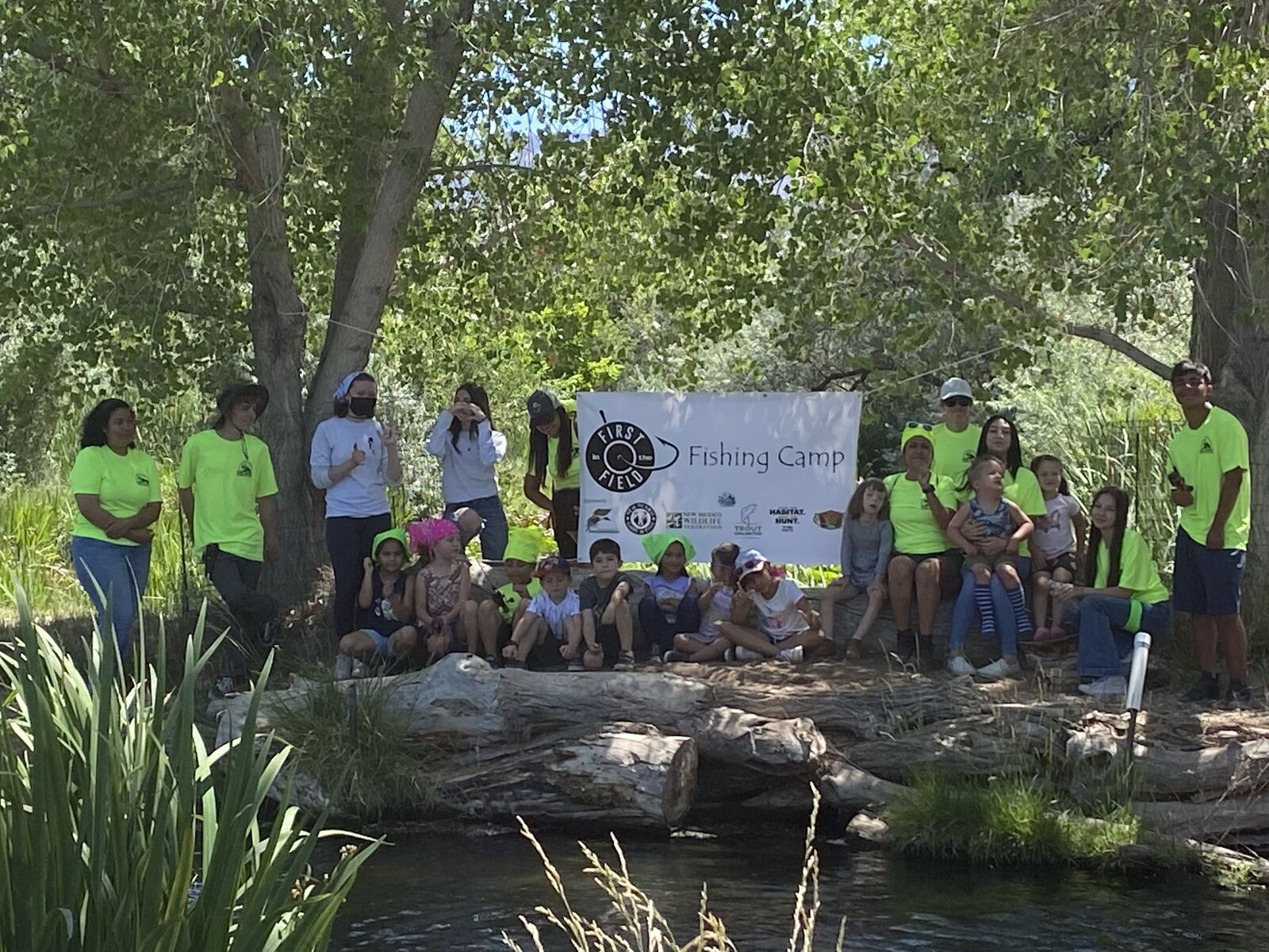 Kids and volunteers gather along a log by the pond under the Fishing Camp banner, in the shade of the cottonwoods.