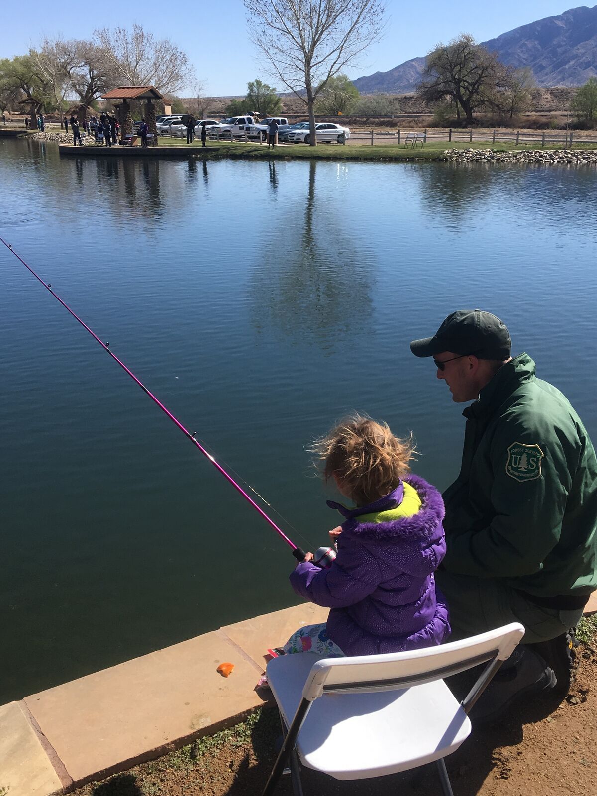 Una niña con chamarra morada pesca a la orilla de un estanque junto a un guardabosques.