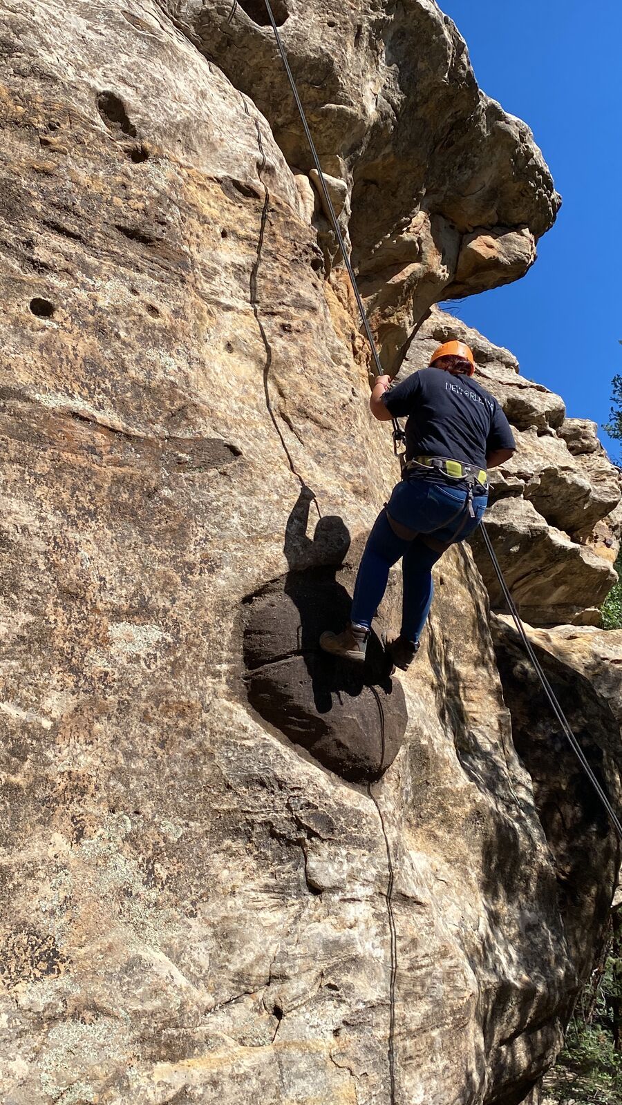 Una persona con casco naranja y arnés sube por una pared de arenisca llena de huecos, atada a una cuerda y de espaldas, bajo un cielo azul intenso.