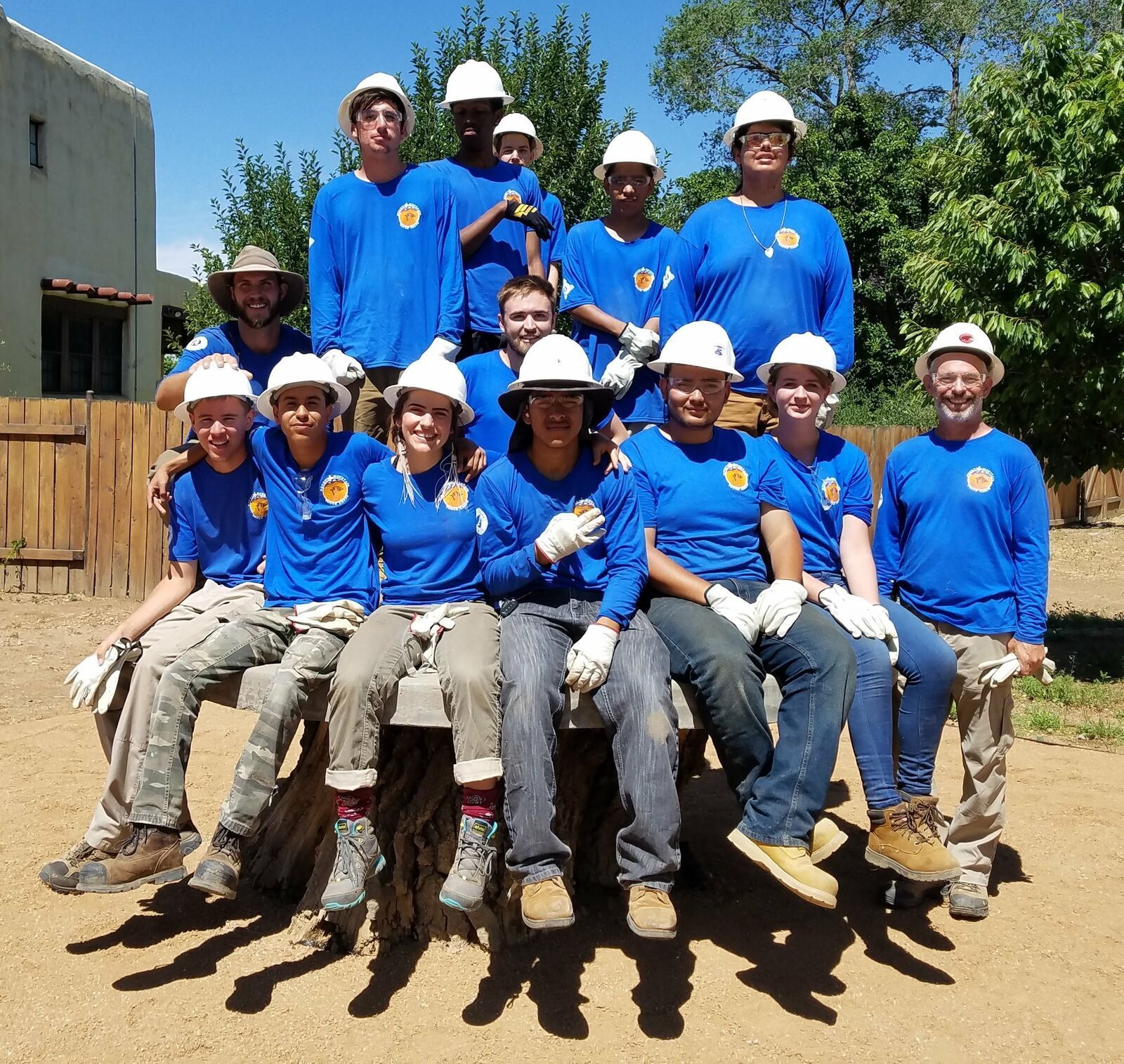 A crew in matching blue shirts, white hard hats and work gloves sits together on a log bench.