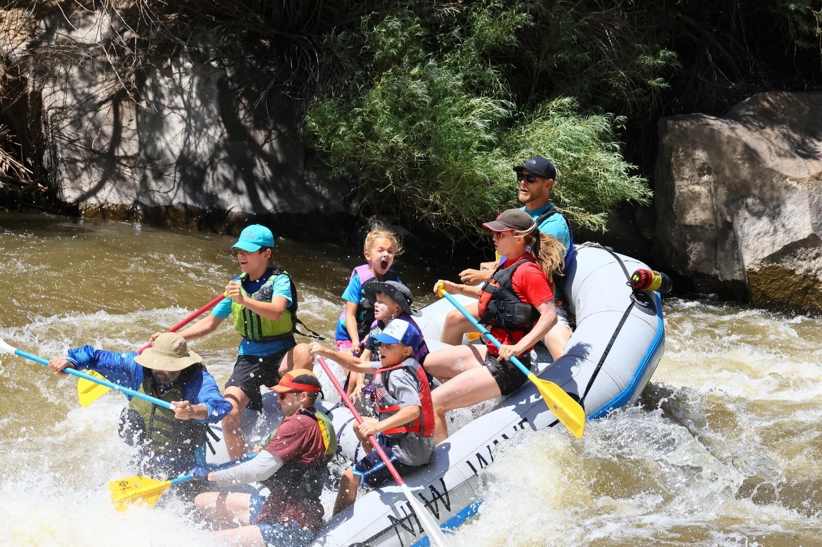 A raft full of kids and grown-ups in life jackets paddles hard through brown whitewater, one girl shouting with excitement.