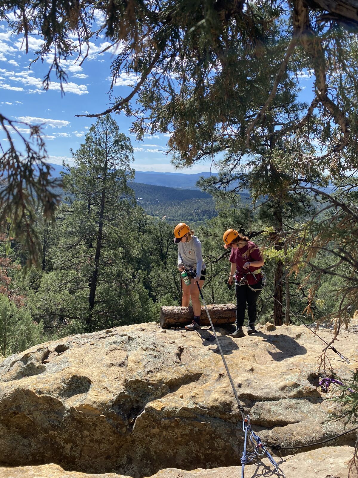 Two climbers in orange helmets on top of a sandstone rock, with pine-covered hills rolling away to the mountains.