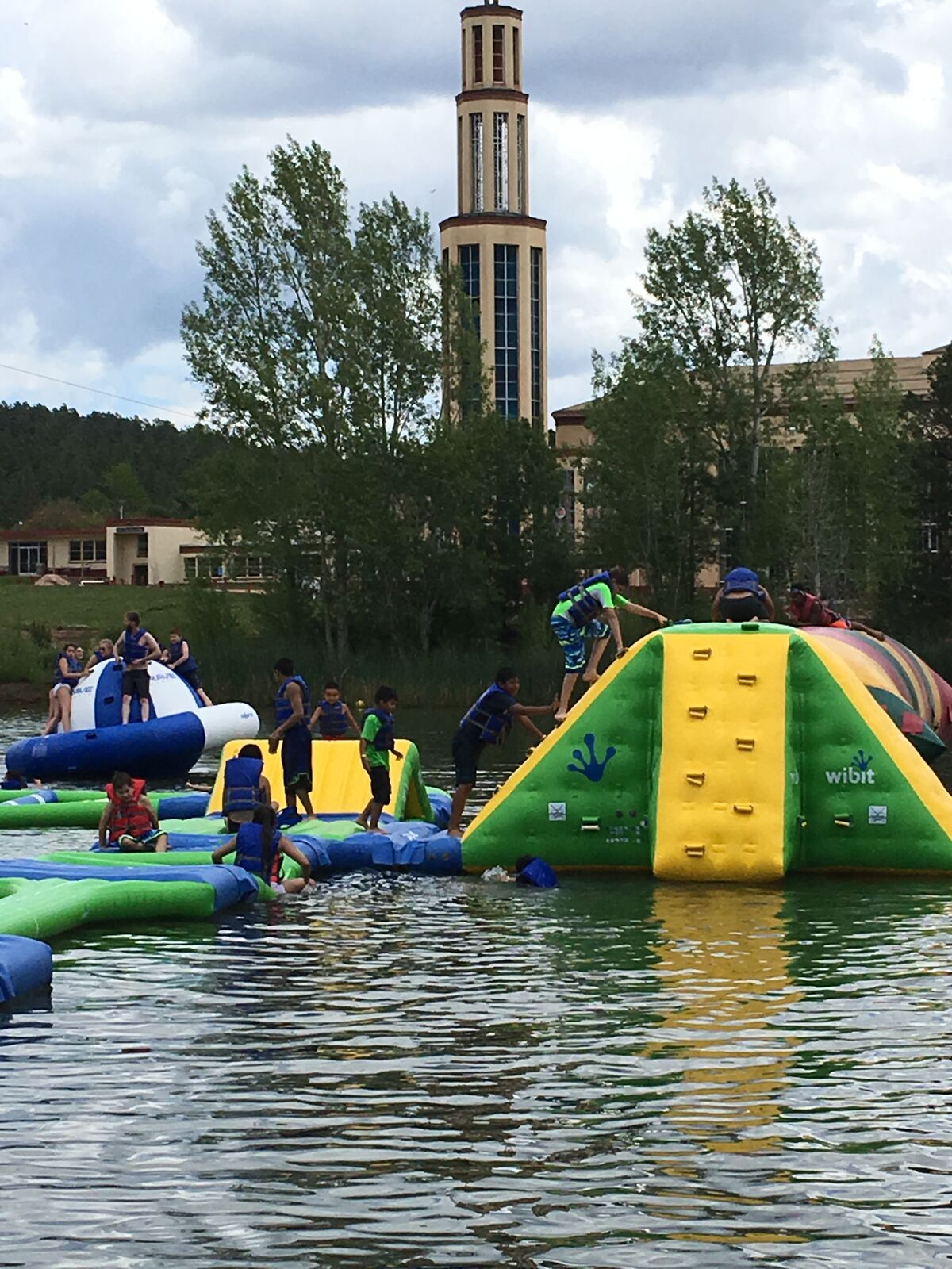 Kids clamber over a bright inflatable water park on the lake at Glorieta, the camp's tall tower rising behind the trees.