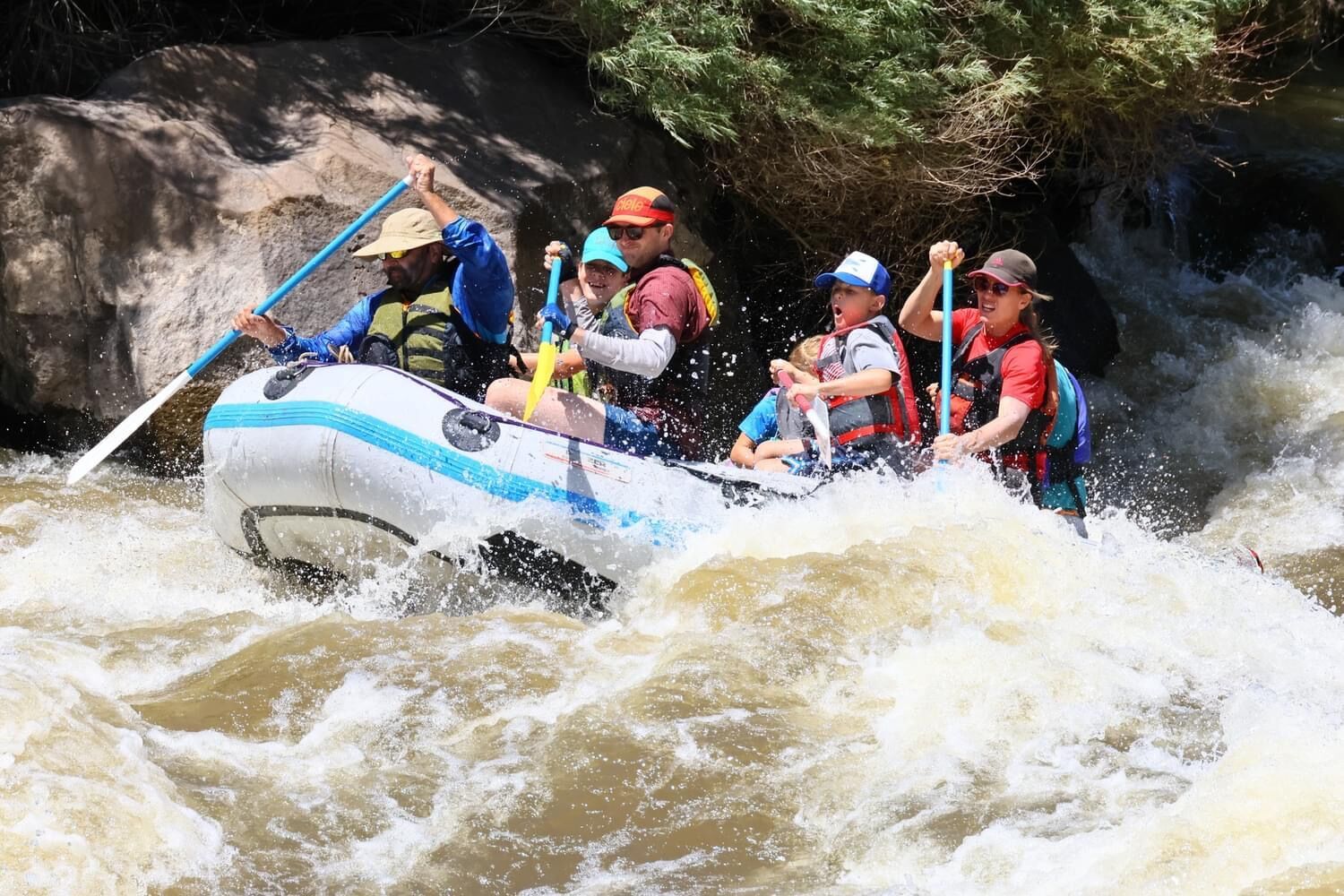 A raft of paddlers in life jackets punches through a big wave in a burst of spray.