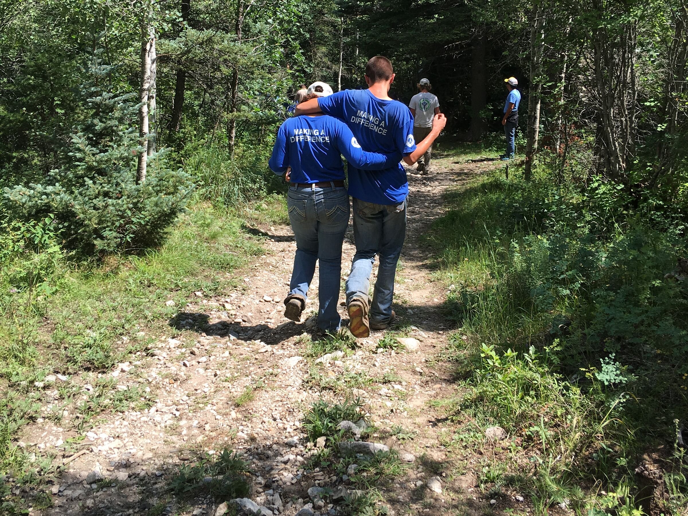 Two people in blue walk a narrow trail into a green forest, seen from behind.