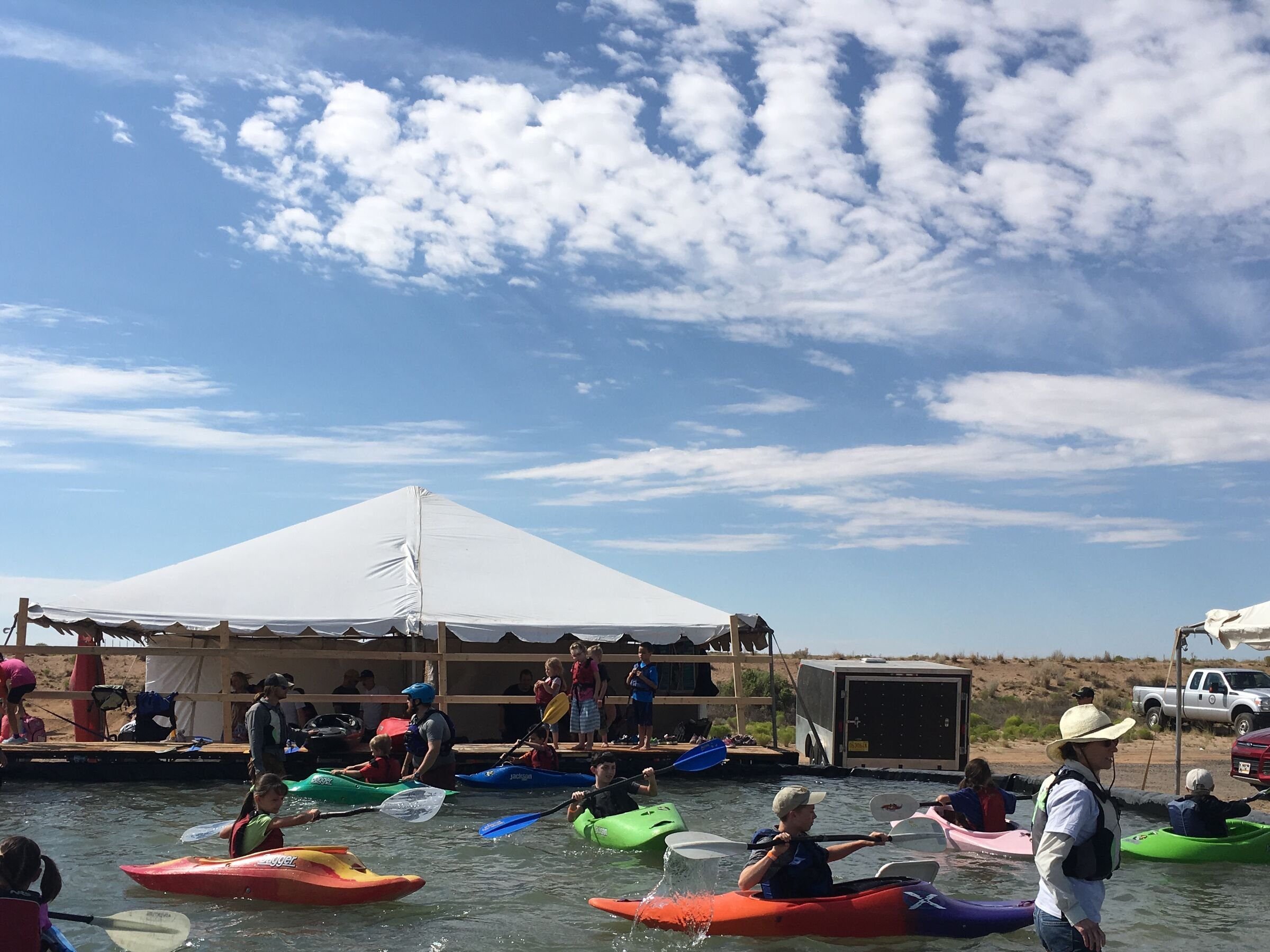 Kids in bright kayaks paddle a shallow pool under a big sky, a white event tent behind.